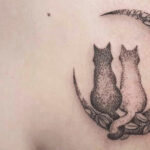 Un tatuaggio sulla parte superiore del braccio raffigurante due gatti seduti su una falce di luna, ornata con motivi floreali.