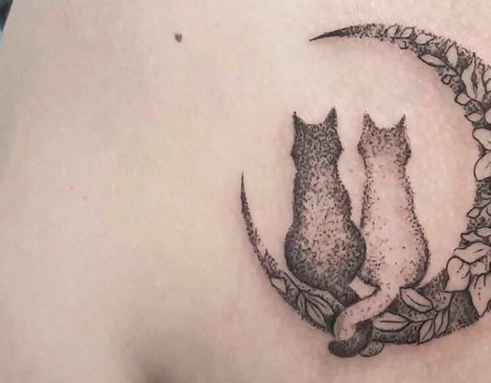 Un tatuaggio sulla parte superiore del braccio raffigurante due gatti seduti su una falce di luna, ornata con motivi floreali.