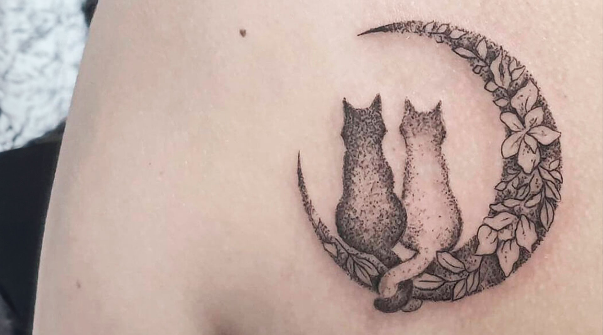 Un tatuaggio sulla parte superiore del braccio raffigurante due gatti seduti su una falce di luna, ornata con motivi floreali.